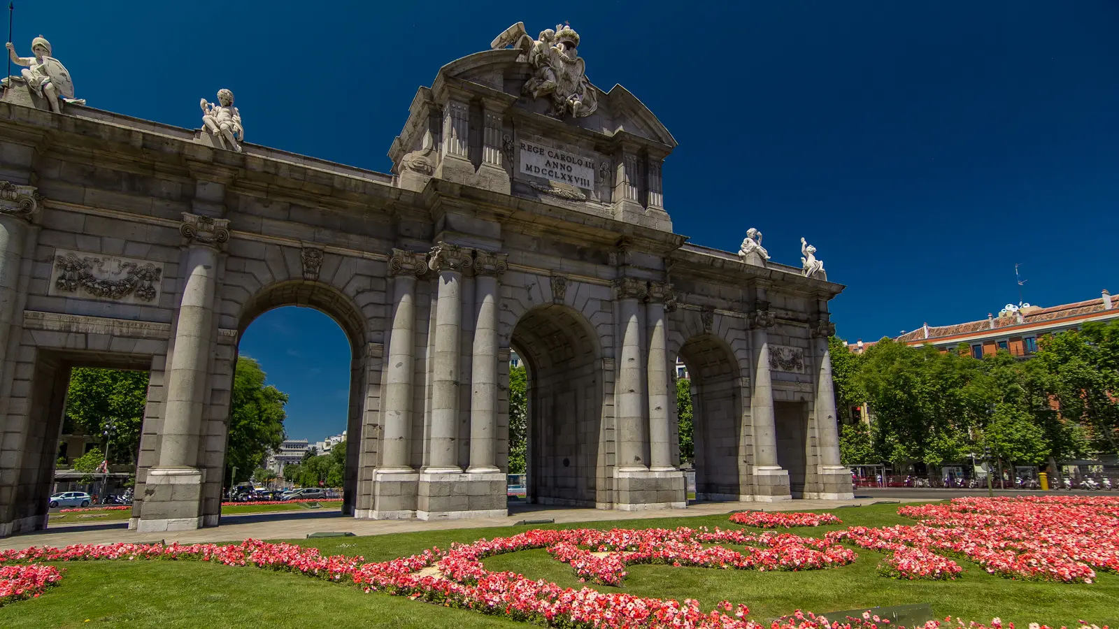 Side view of the Puerta de Alcalá with red flower beds, showing the full Neoclassical facade and sculptural details