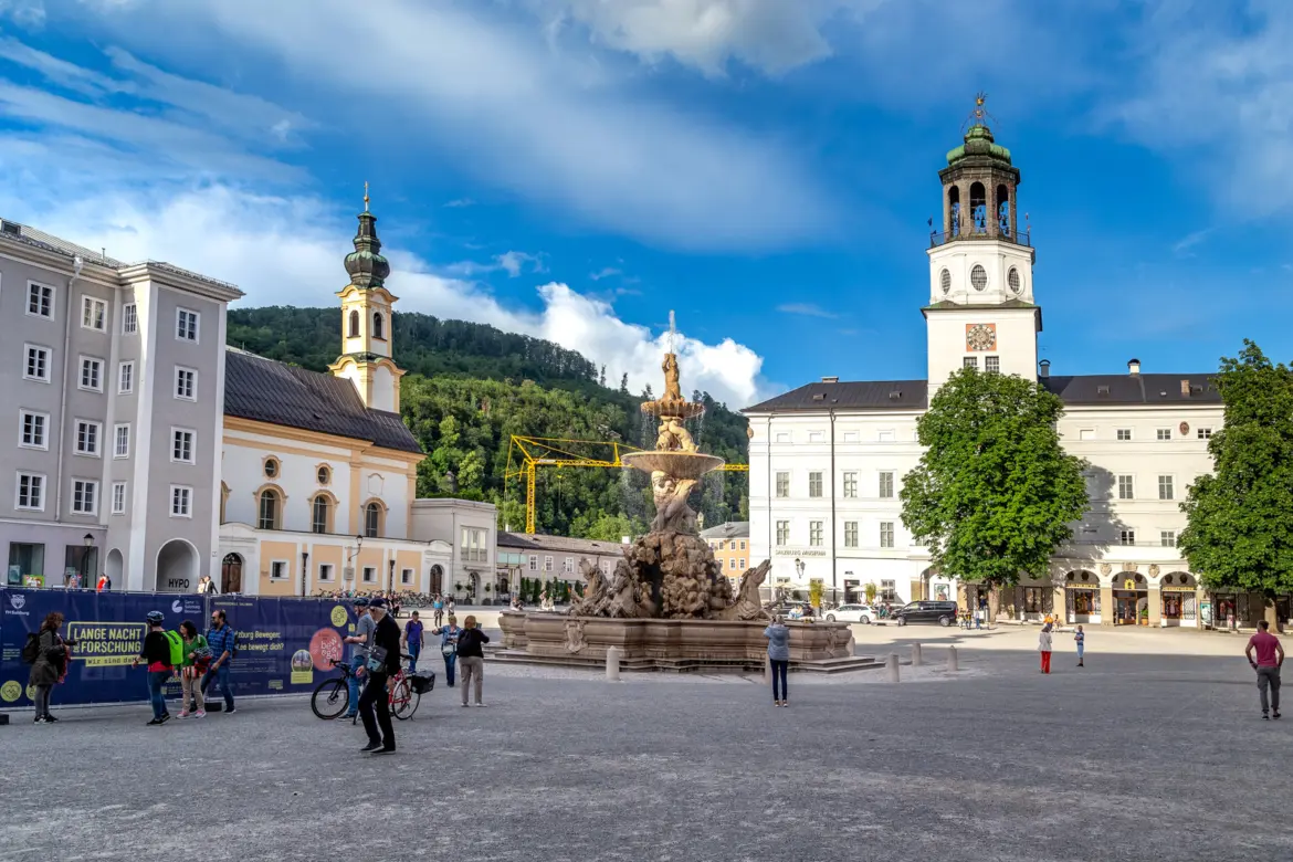 People walking across Residenzplatz on a sunny day in Salzburg with the Residenzbrunnen fountain, Michaelskirche and Glockenspiel tower