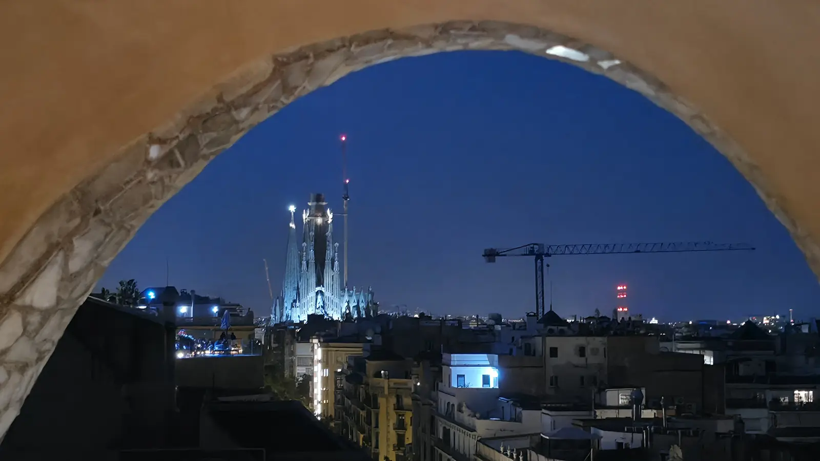The Sagrada Família illuminated at night as seen from the rooftop of La Pedrera (Casa Milà) in Barcelona, its spires glowing against the dark sky above the city's rooftops