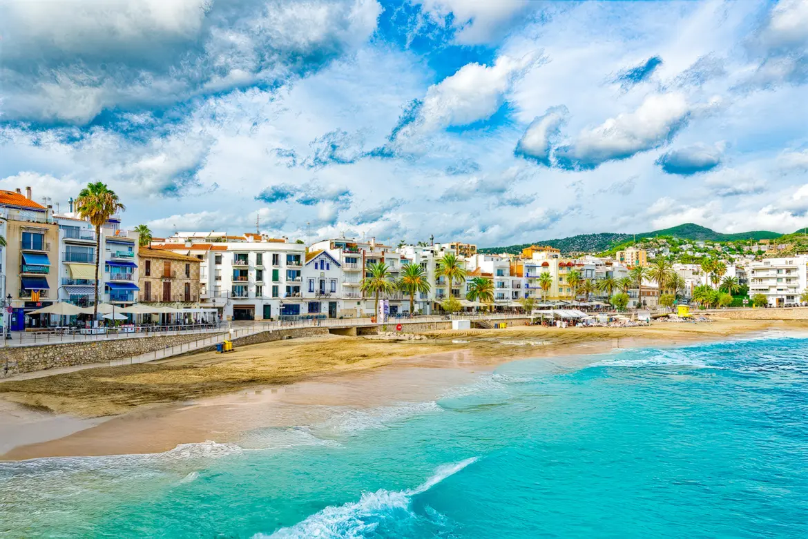 Sitges on the Mediterranean coast of Catalonia, with its iconic church perched above golden sand, whitewashed buildings cascading toward the sea, and gentle waves catching the sunlight