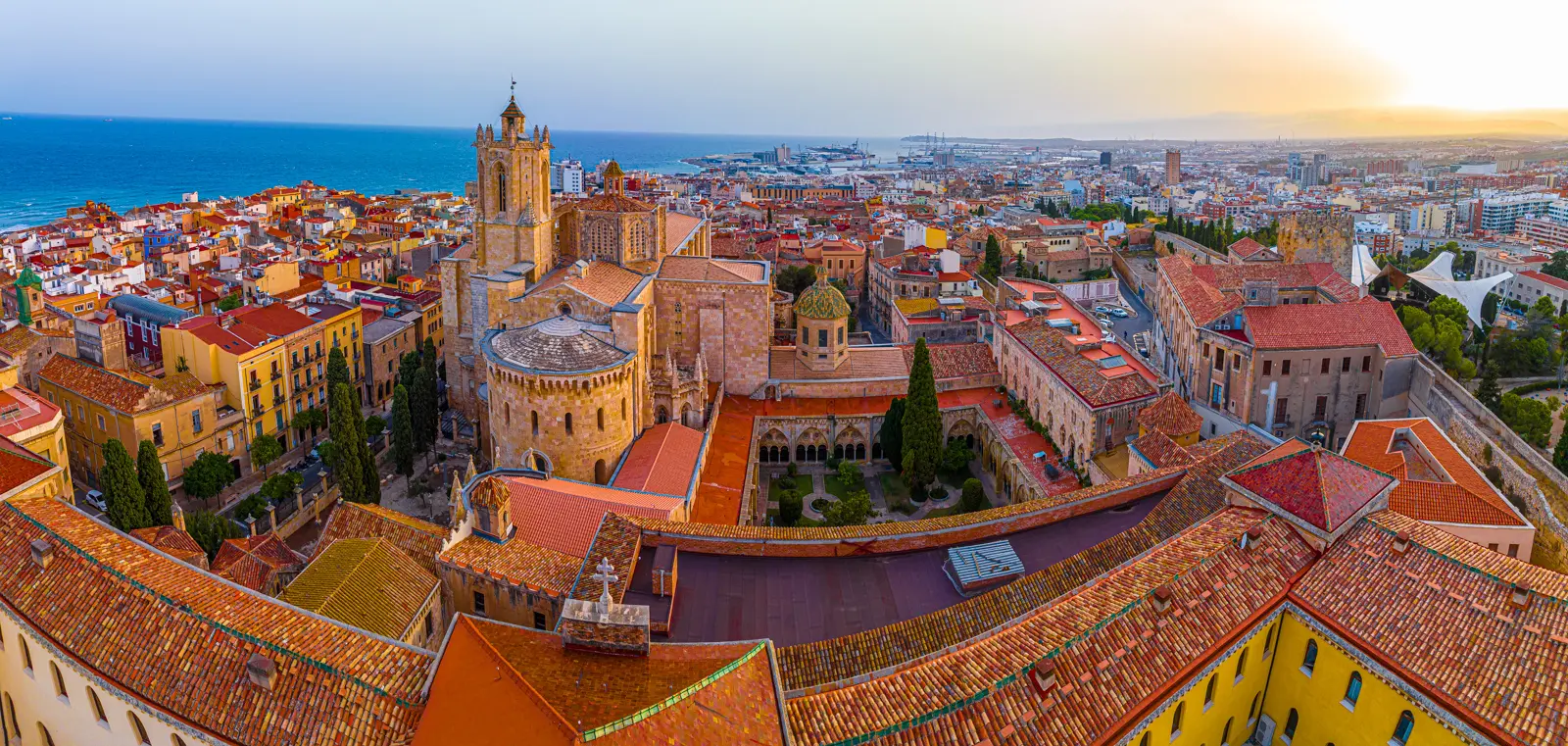 A sweeping panoramic view of Tarragona's golden beach curving gracefully along turquoise Mediterranean waters, with the ancient city rising behind the coastline
