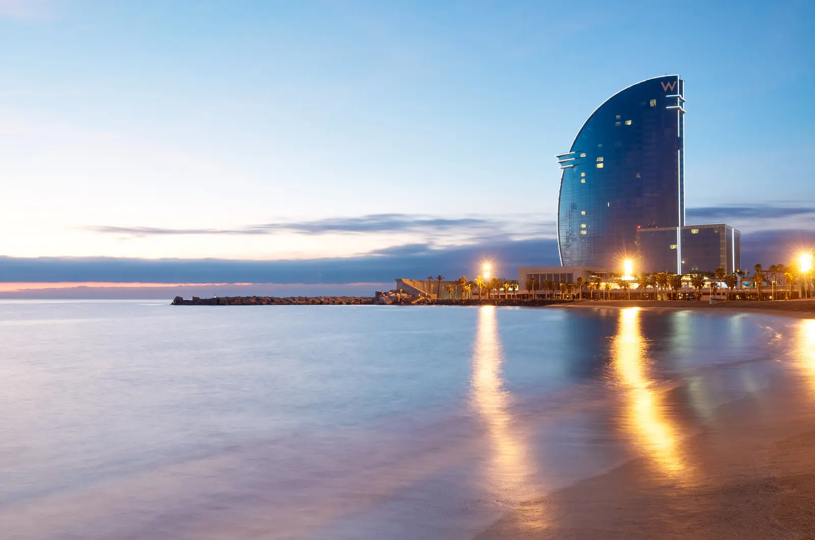 The sail-shaped W Barcelona Hotel (Hotel Vela) at the edge of Barceloneta Beach, reflecting gold and rose sunset light across the glass façade