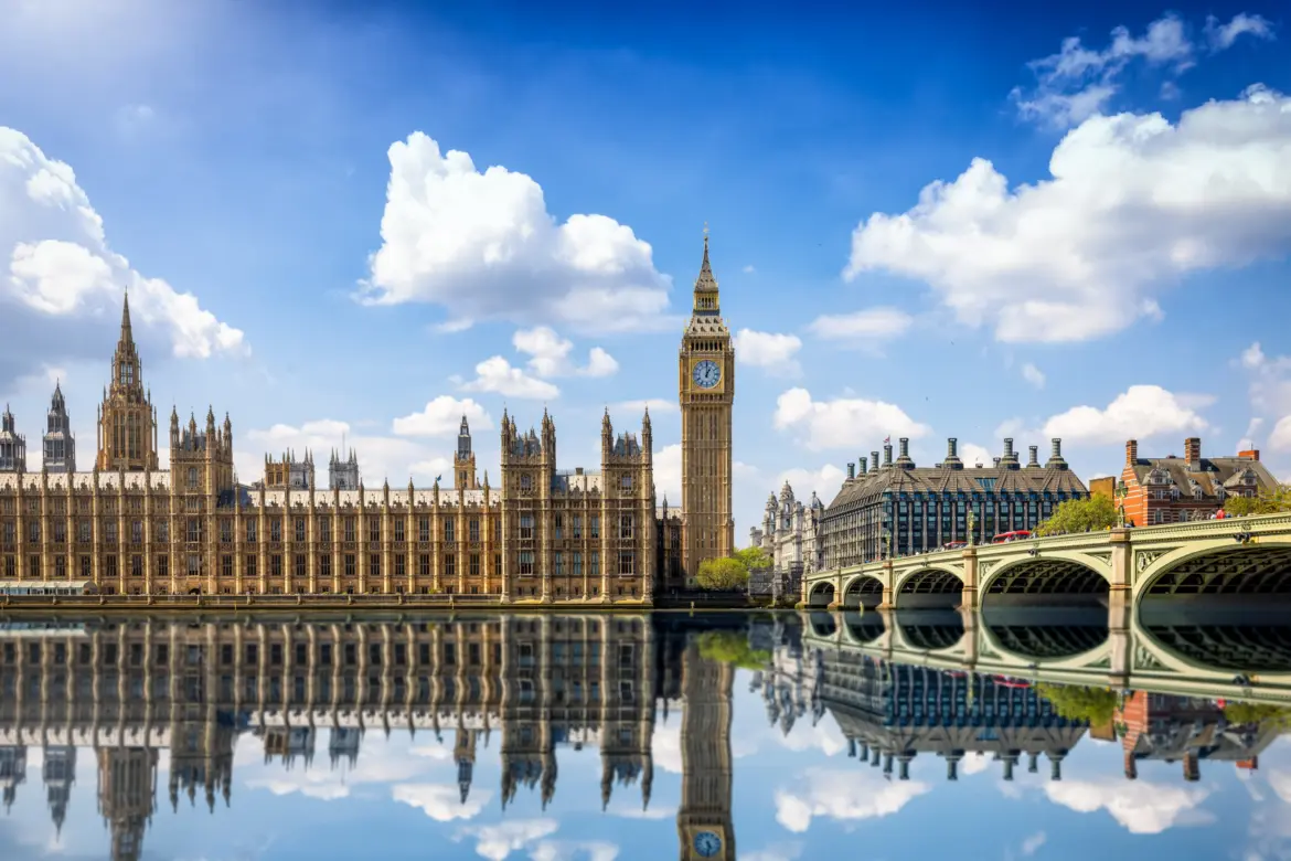 The Elizabeth Tower (Big Ben), the Palace of Westminster, and Westminster Bridge reflected in the still waters of the River Thames in London under a bright sky with dramatic clouds