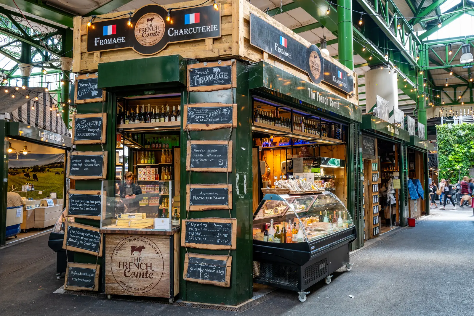 The French Comté cheese-and-charcuterie stall at Borough Market, painted green with hand-lettered chalkboard signs reading Fromage, Charcuterie, Saucissons, and Comté Cheese 12, 18, 24 and 36 month