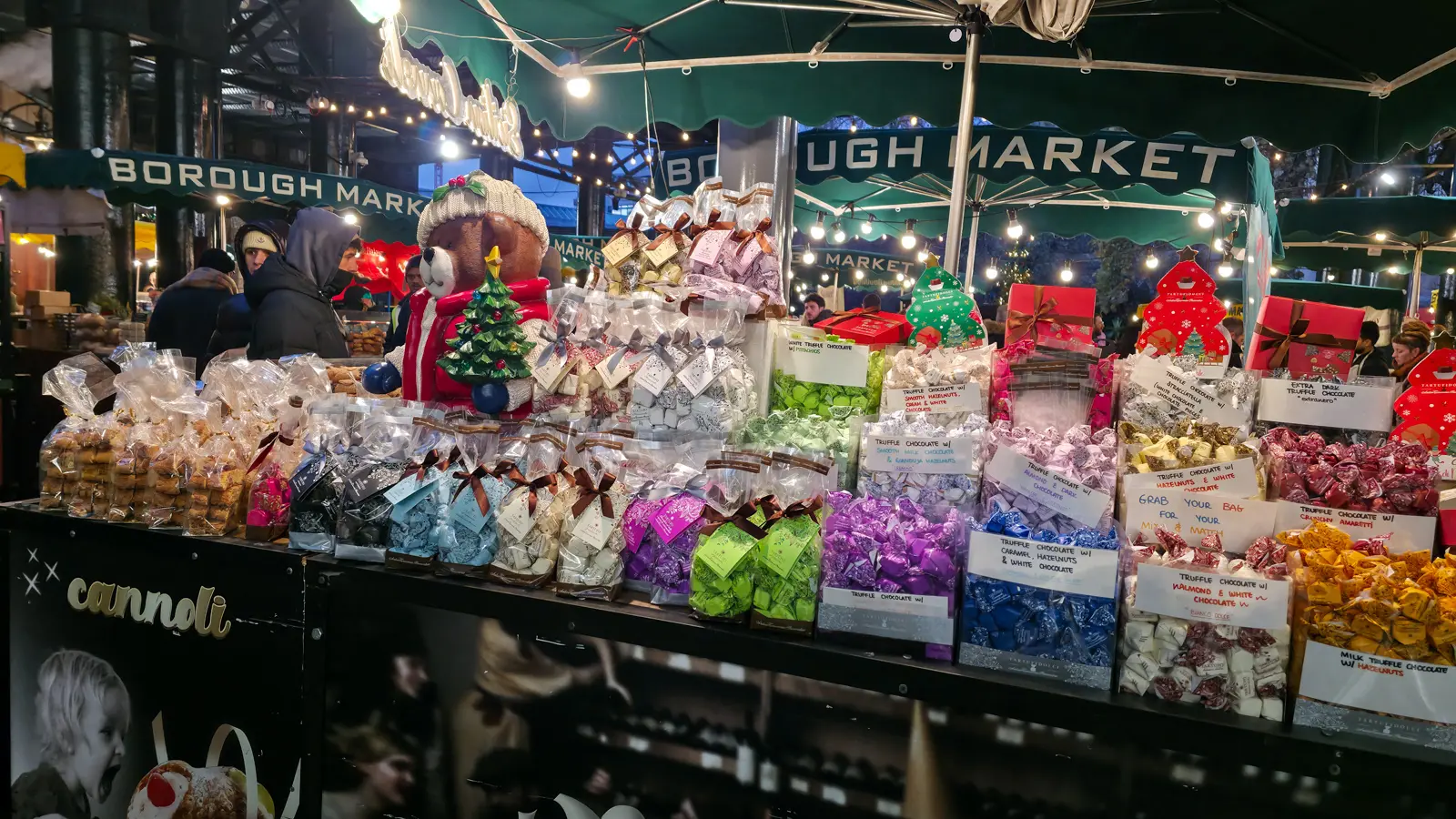 Italian truffle chocolate stall at Borough Market's Christmas market with hand-lettered signs for flavours including tiramisu, hazelnut and white chocolate, caramel hazelnut, amaretti, and extra dark, plus festive packaging and a teddy bear holding a Christmas tree