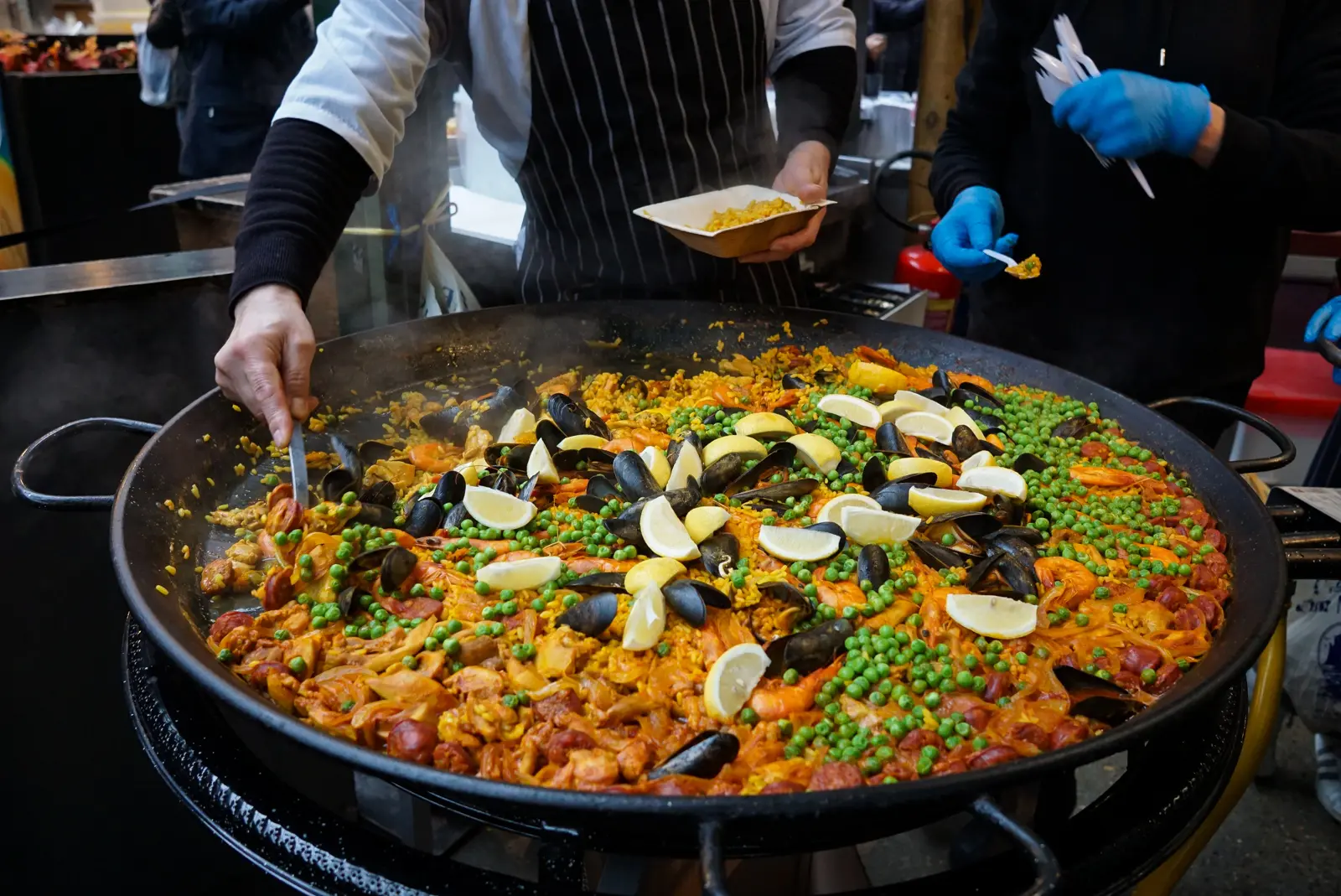 A vendor in a striped apron serving paella into a wooden takeaway tray from a large black paella pan filled with saffron rice, mussels, prawns, chorizo, peas, and lemon wedges