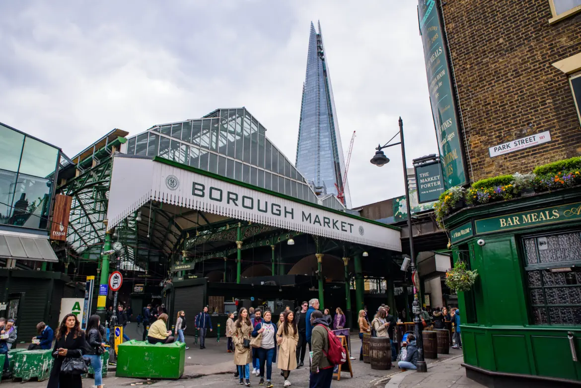 The main Borough Market entrance on Park Street in Southwark beneath the green wrought-iron Borough Market sign, with The Shard rising directly behind the Victorian iron-and-glass hall and The Market Porter pub on the right