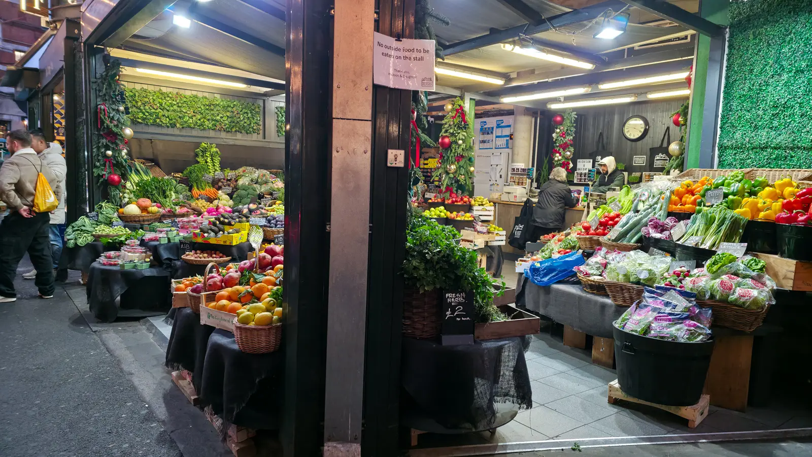 A Borough Market produce stall decorated with Christmas garlands and gold baubles, with woven baskets of pomegranates, oranges, and persimmons on a black draped table, and a wider counter of bell peppers, tomatoes, lettuce, and lemons behind