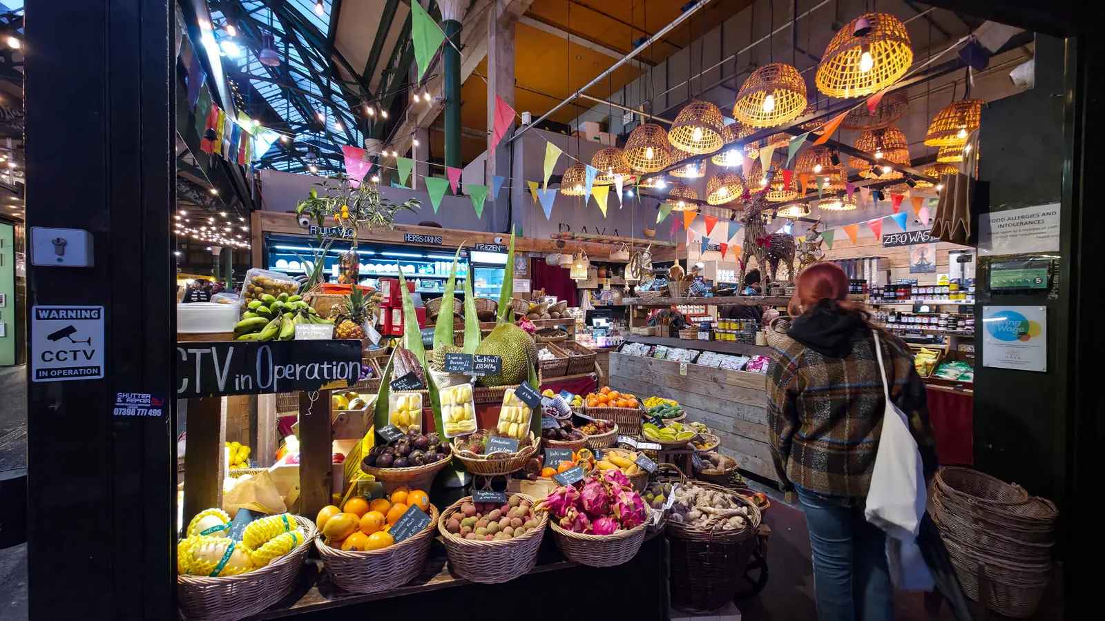 Tropical fruit stall at Borough Market under woven rattan pendant lights and colourful bunting, with a centre display of jackfruit, dragon fruit, lychees, rambutans, baby bananas, ginger, and citrus in wicker baskets