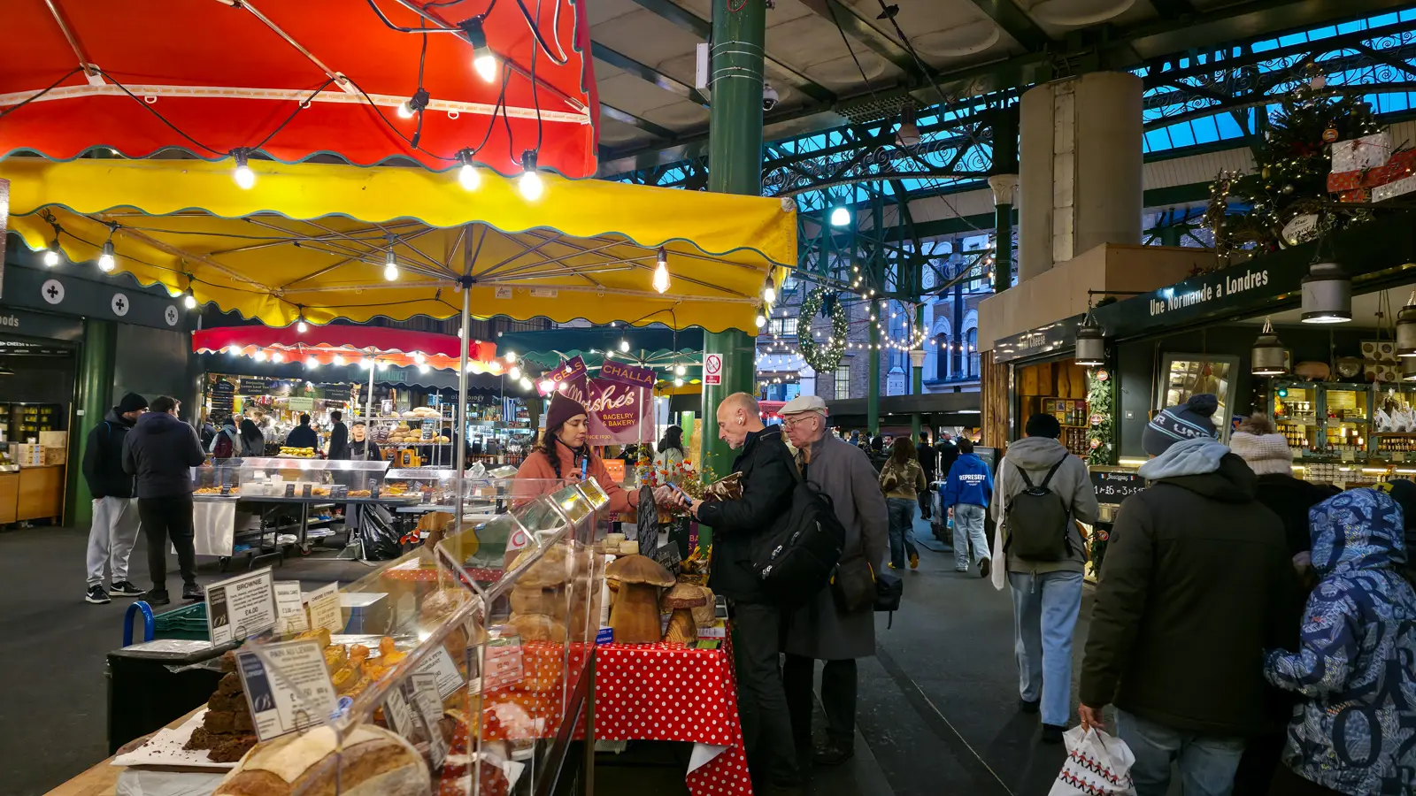 The interior of Borough Market's Victorian iron-and-glass hall in December, with Moishe's Bagelry banner visible above a foreground stall under red-and-yellow striped umbrellas, Christmas string lights overhead, and an Une Normande à Londres cheese counter on the right