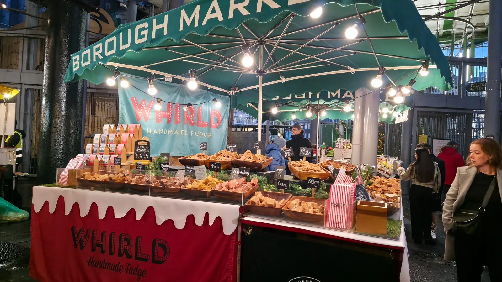 The Whirld Handmade Fudge stall at Borough Market in December under a green Borough Market parasol, displaying wooden bowls of fudge in flavours including chocolate orange, mango, raspberry, white chocolate, gingerbread latte, hot honey, salted caramel, and Baileys, with a red-and-white drip-painted front sign