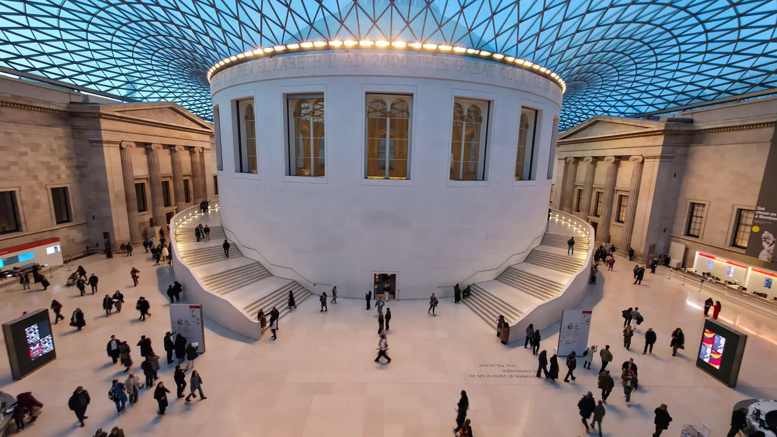 The Great Court of the British Museum in London with its tessellated glass roof above the central Reading Room rotunda, marble floor, and visitors moving through the vast covered space