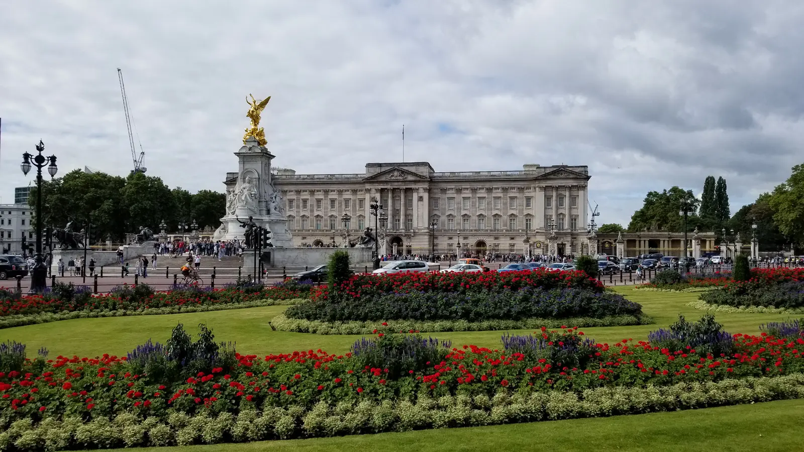 Buckingham Palace in London framed by the Memorial Gardens with red and purple summer flower beds in the foreground, the gilded Queen Victoria Memorial to the left, and overcast skies