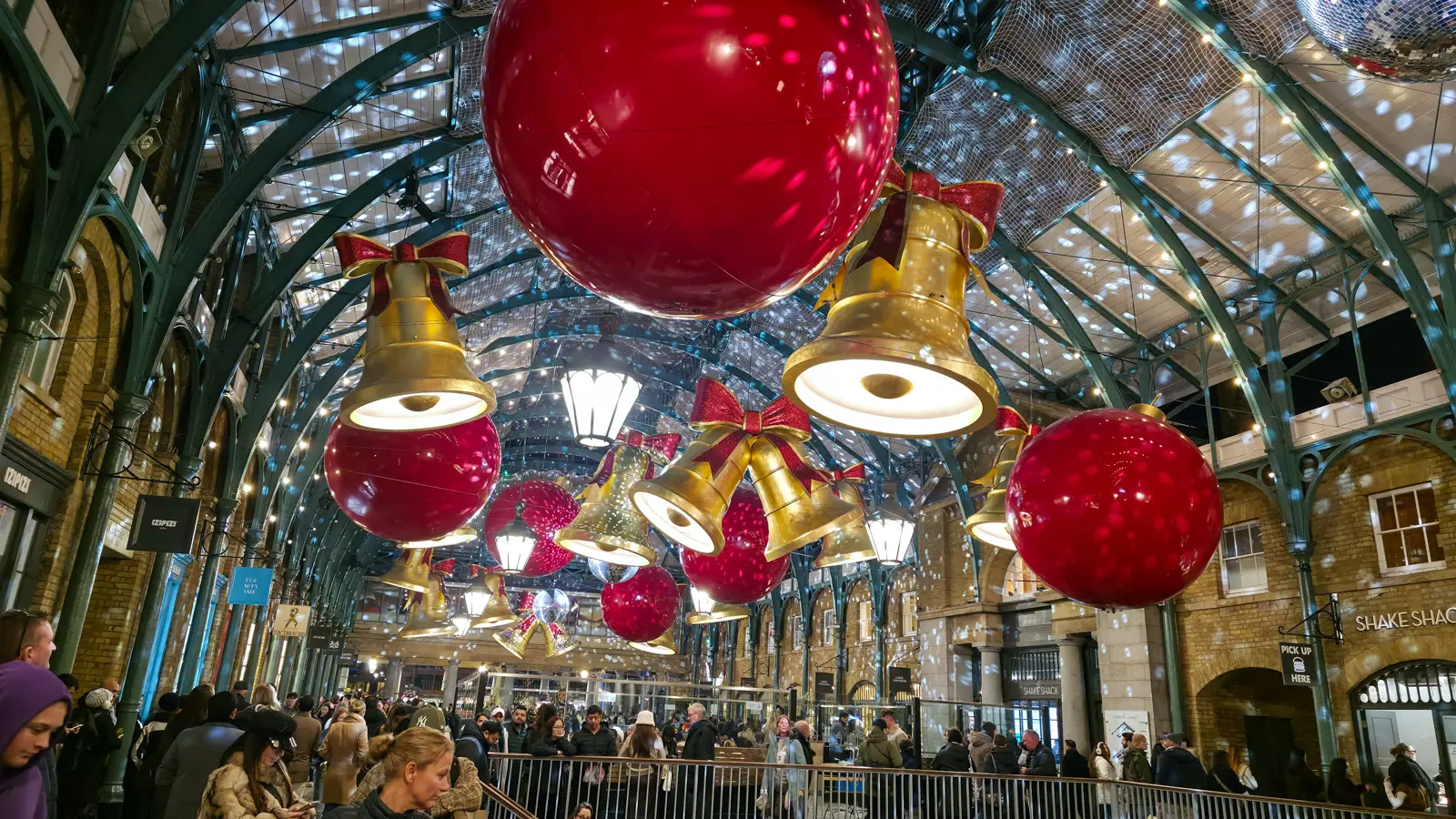 Giant golden bells with red ribbons and oversized red baubles suspended from the ironwork ceiling of Covent Garden Market in London during the Christmas season, with crowds visible below