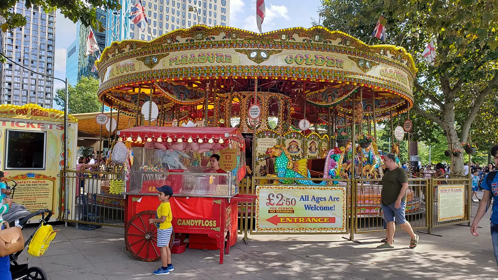 A vintage-style fairground carousel with hand-painted sea-creature mounts and an attendant candyfloss cart on the South Bank, set up near Jubilee Gardens and the Royal Festival Hall. Rides are listed at £2.50 per person on the entrance sign, and family-friendly attractions appear along this stretch throughout the warmer months