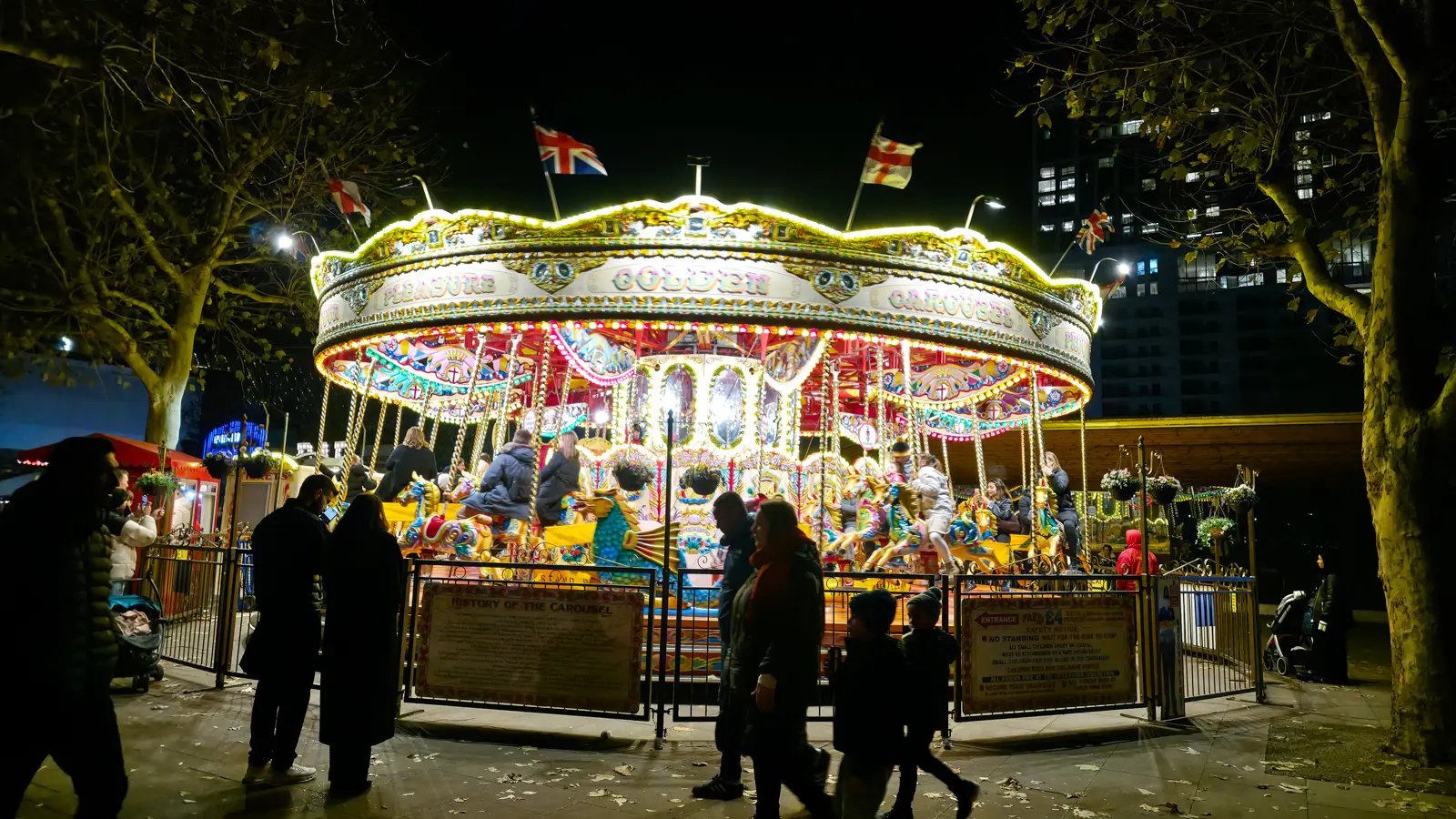 The Golden Carousel sets up beside Jubilee Gardens during the Winter Market and runs into the evening, the canopy strung with bulbs against the December dark. The history of the carousel is posted on the entrance gate and tickets are sold from the booth on the left. Cash only