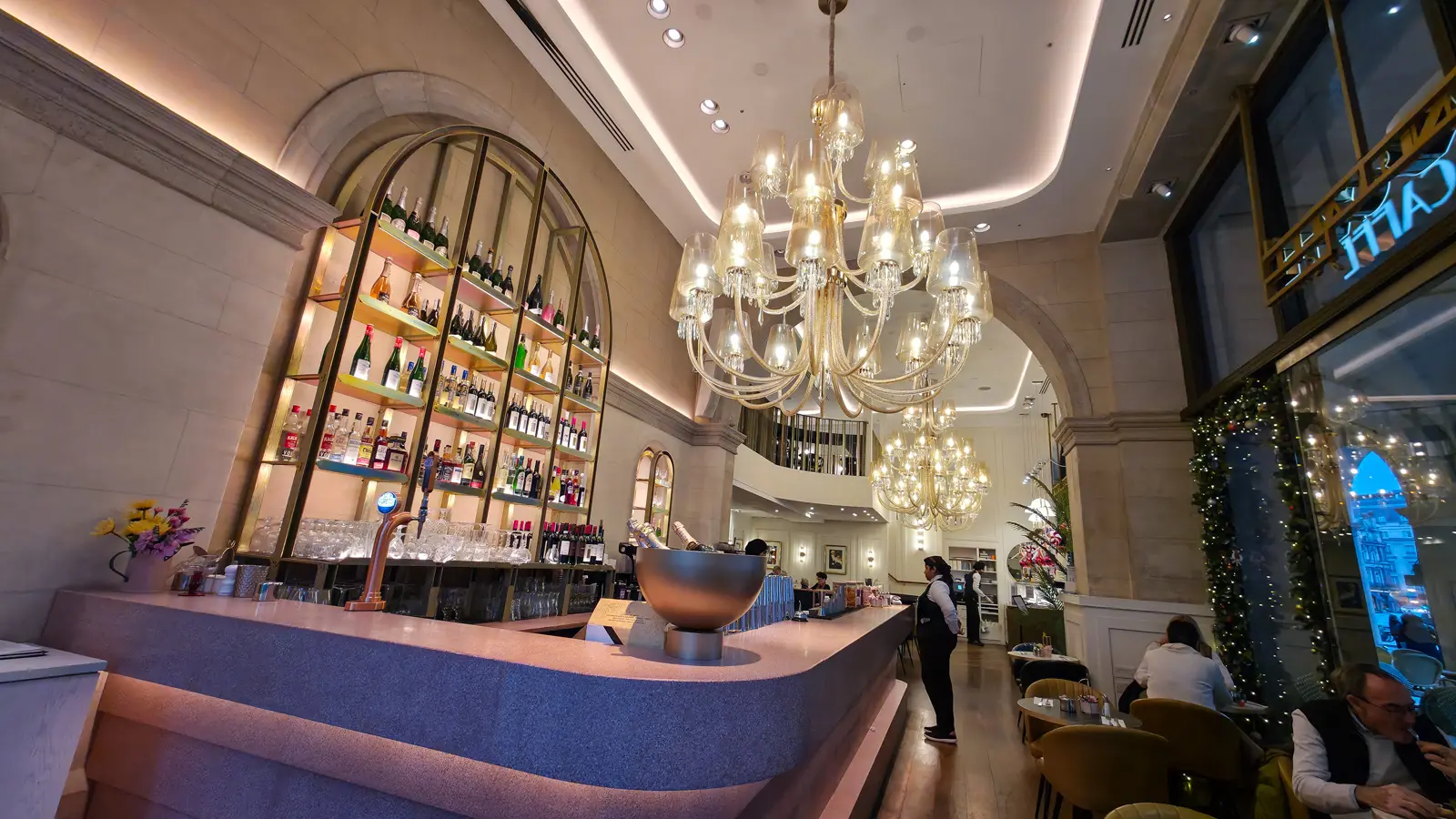 The bar at The Grand by Concerto on the Strand, with a pink terrazzo counter, an arched bronze-framed display of wine, champagne, and prosecco bottles behind it, and two enormous crystal chandeliers cascading from the high ceiling