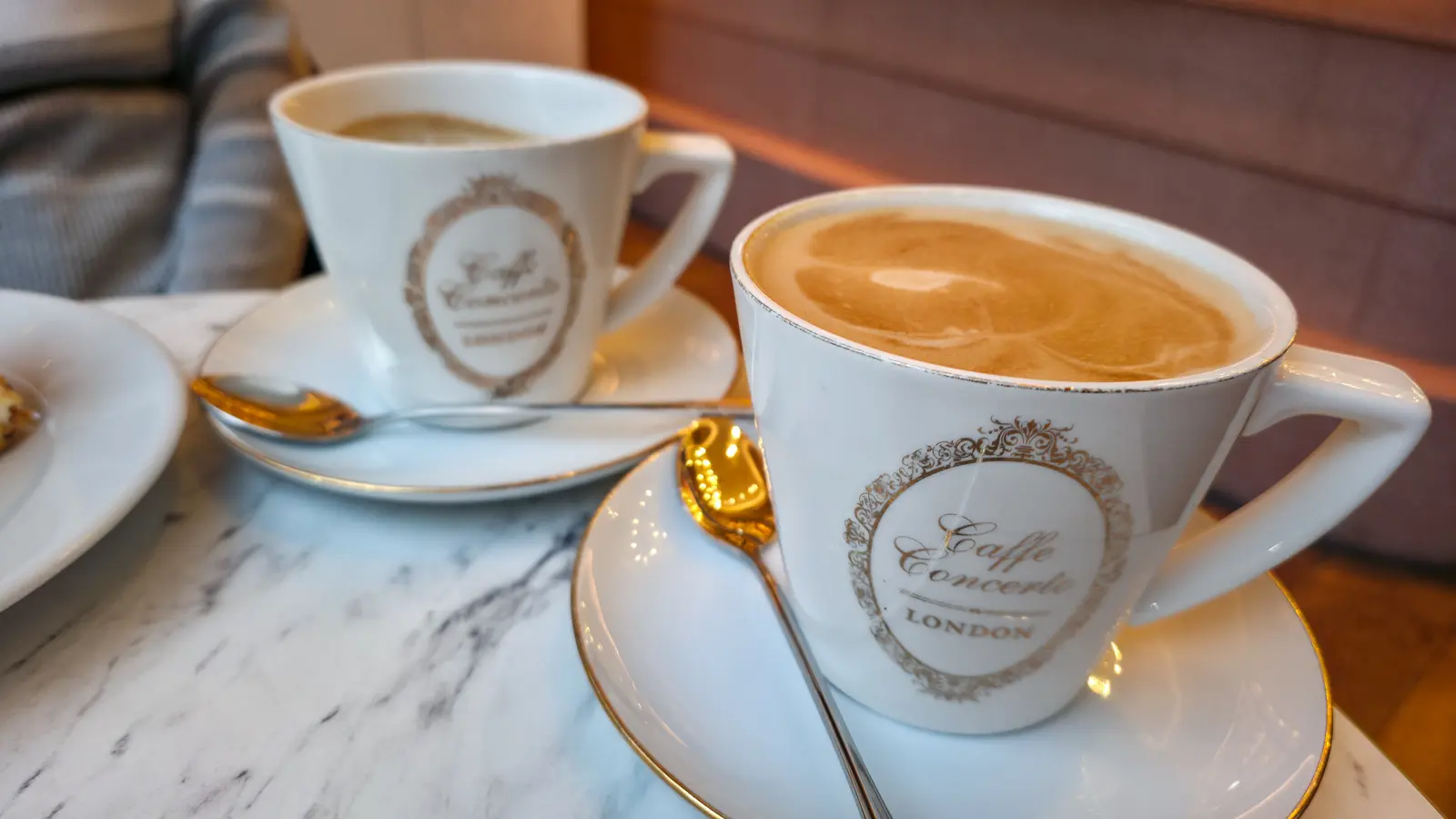 Two cappuccinos in white porcelain Caffè Concerto London cups with gold-rim trim and the chain's ornate gold scrollwork crest, on white saucers with gold-handled spoons, photographed on a marble tabletop