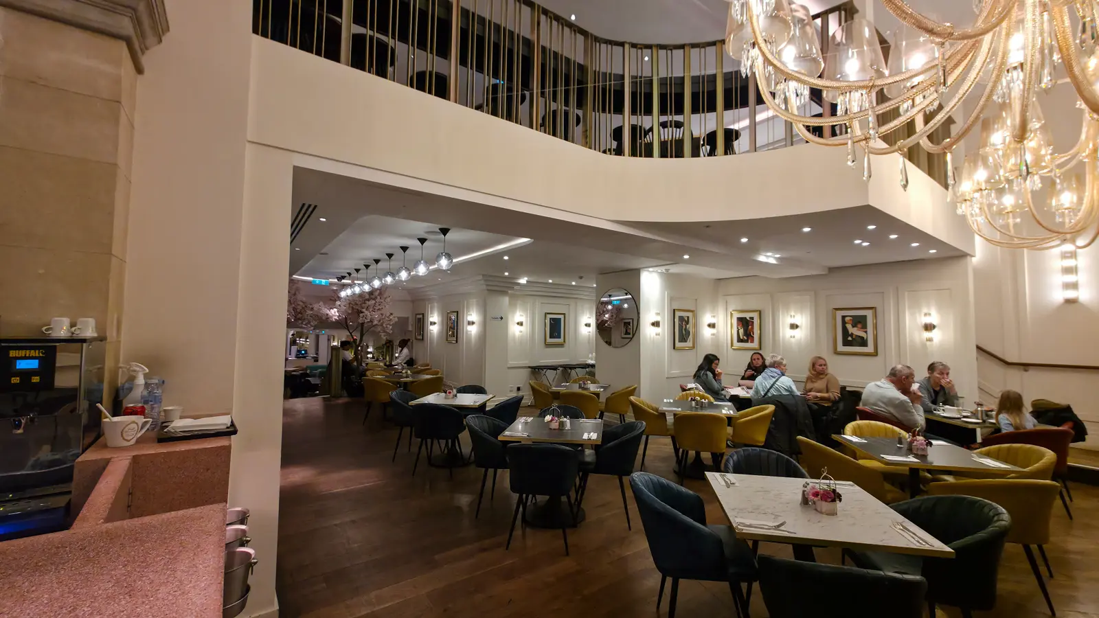The main dining room at The Grand by Concerto on the Strand, looking down a two-storey space with cream walls and decorative cornices, two enormous cascading crystal chandeliers in the foreground, framed portrait paintings along the right wall, and marble tables with dark velvet chairs in yellow and forest green throughout