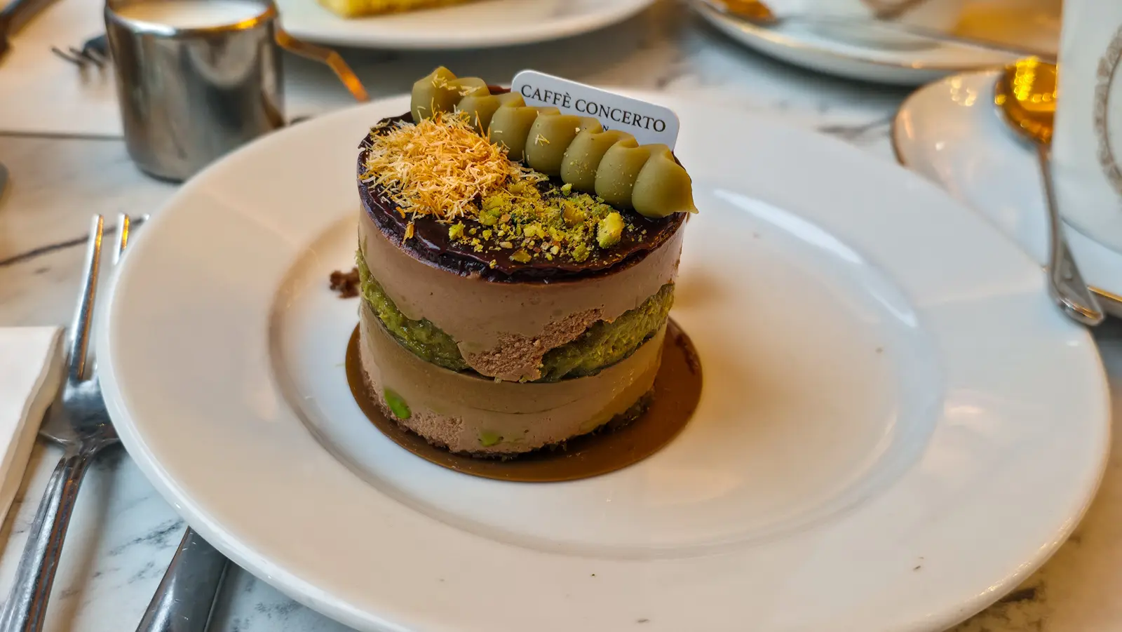 The Dubai chocolate cake at The Grand by Concerto, a small round individual cake about the size of a teacup with visible side layers of chocolate mousse and pistachio sponge, topped with a chocolate ganache disc, shredded golden kataifi pastry, crushed pistachios, and a row of six pistachio cream rosettes holding a small Caffè Concerto card