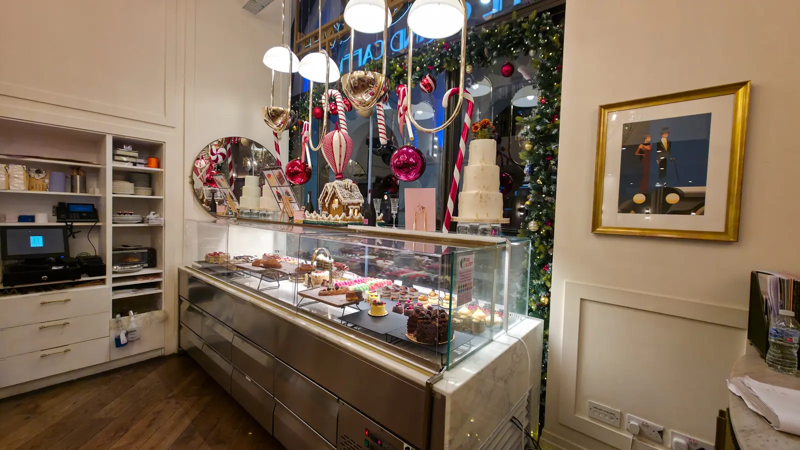 The pastry cabinet at The Grand by Concerto in December, with an architectural gingerbread house, oversized red and white candy cane decorations hanging from the ceiling, pink and red mirror baubles, and a three-tier white wedding cake on top, with the actual pastry case below holding rows of macarons, fruit tarts, opera cakes, and individual gateaux