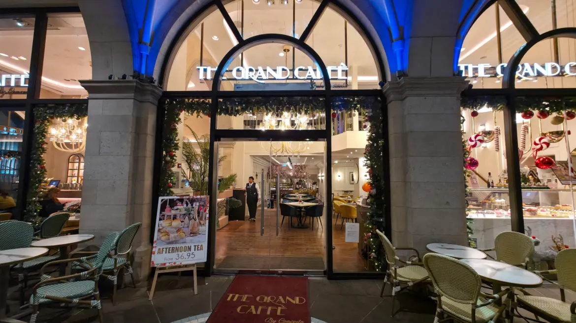 The entrance to The Grand by Concerto on the Strand at night, with the preserved Victorian arched façade lit blue, the THE GRAND CAFFÈ signage above the doors, Christmas garlands wrapping the arches, an afternoon tea easel sign reading from £36.95, and the warm interior visible through the open doors with the chandeliers, cherry blossom trees, and dining room beyond