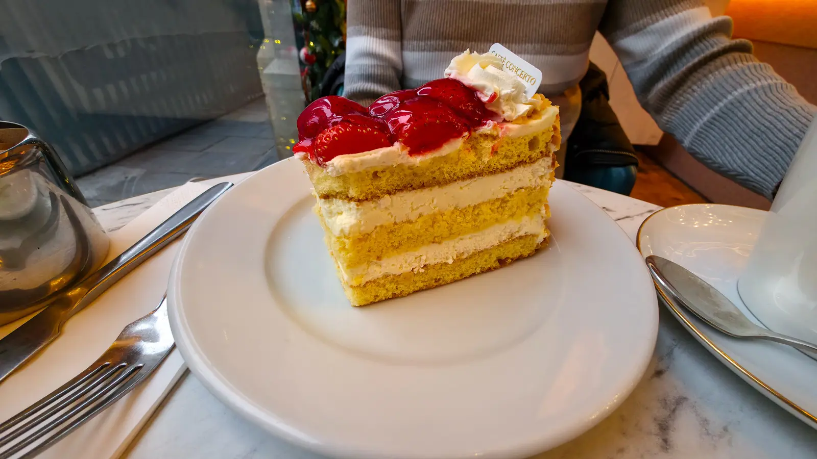 A slice of strawberry gateau at The Grand by Concerto, three layers of pale yellow vanilla sponge with cream filling between each layer, topped with glazed strawberries and a piped cream rosette holding a small white Caffè Concerto card, on a white plate against a marble tabletop