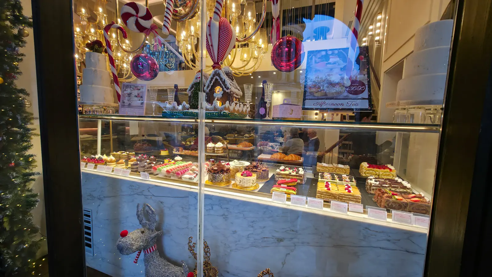 The Caffè Concerto pastry vitrine viewed from the Strand at night, with the warm interior chandeliers lighting the three-tier white wedding cake at the top of the display, the gingerbread house and oversized red and white candy canes at centre, hanging pink and red baubles, a row of macarons and gateaux in the cabinet below, and silver and gold wire reindeer figures visible in the foreground reflection