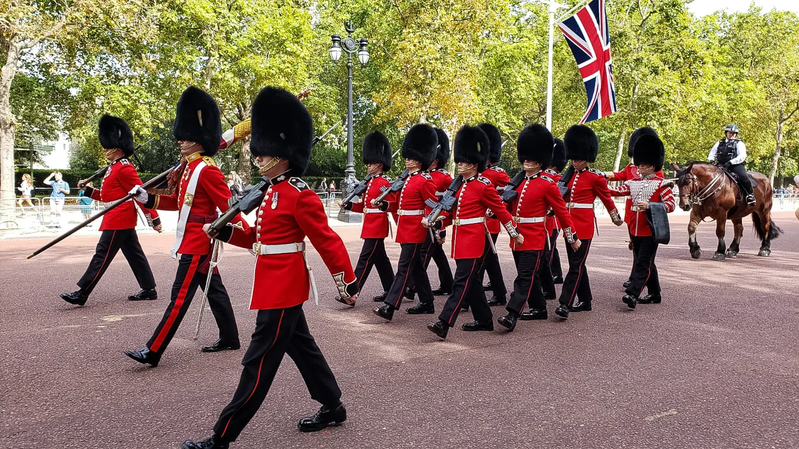 A column of King's Guards in red tunics and bearskin hats marching down The Mall in London with rifles shouldered, a mounted police officer alongside, and a Union Jack flag overhead