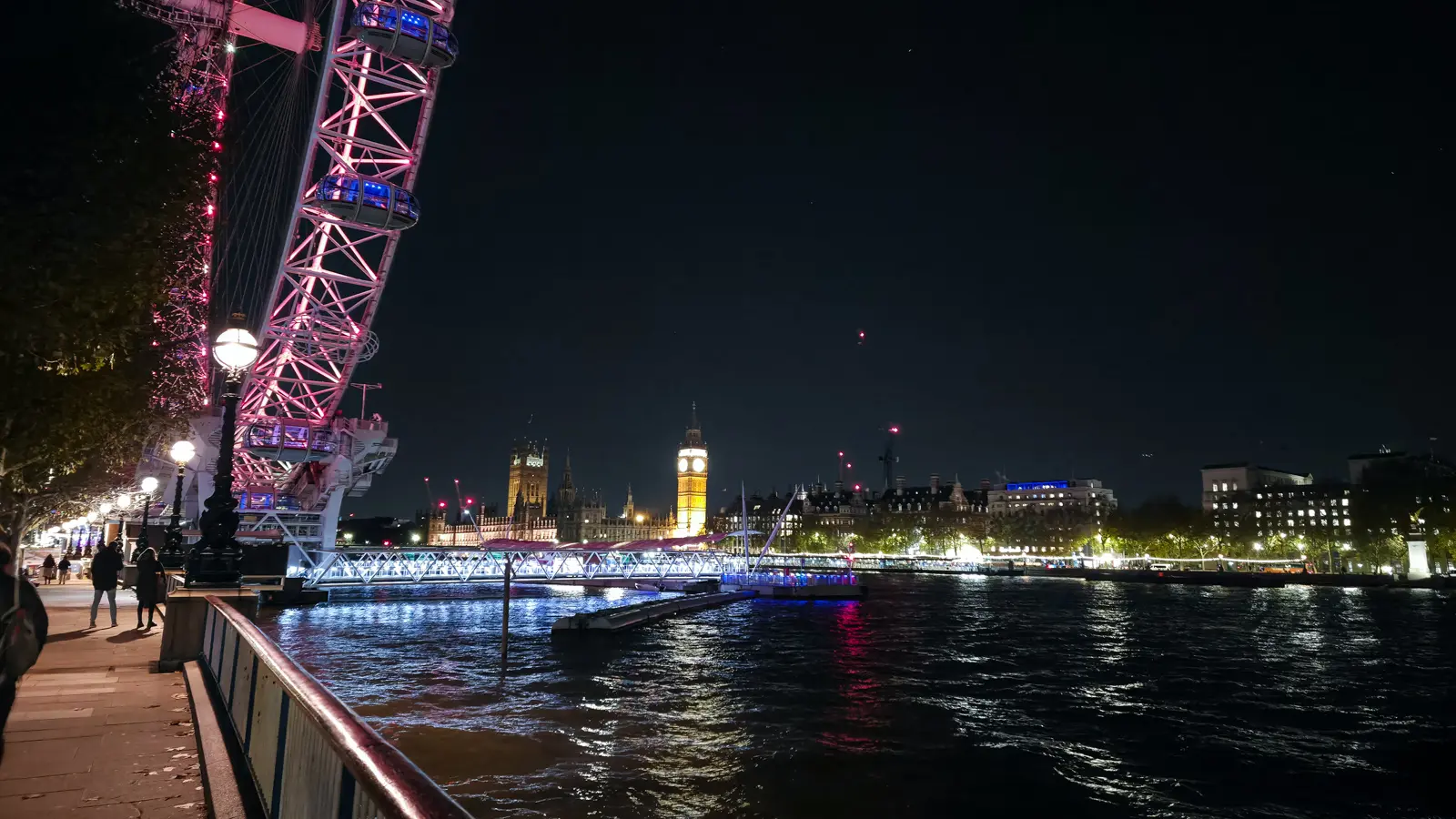 Night view from the South Bank in London with the illuminated London Eye in pink and white on the left, Big Ben glowing golden across the Thames, and the river reflecting both