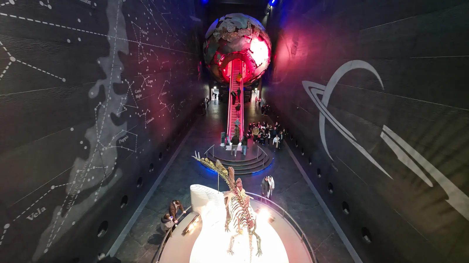 The Earth Hall atrium at the Natural History Museum, with the red-lit escalator rising through the centre of the glowing metallic globe of the Earth, Sophie the stegosaurus skeleton illuminated on a circular plinth at the foot of the escalator, and constellation maps engraved on the dark slate walls of the atrium