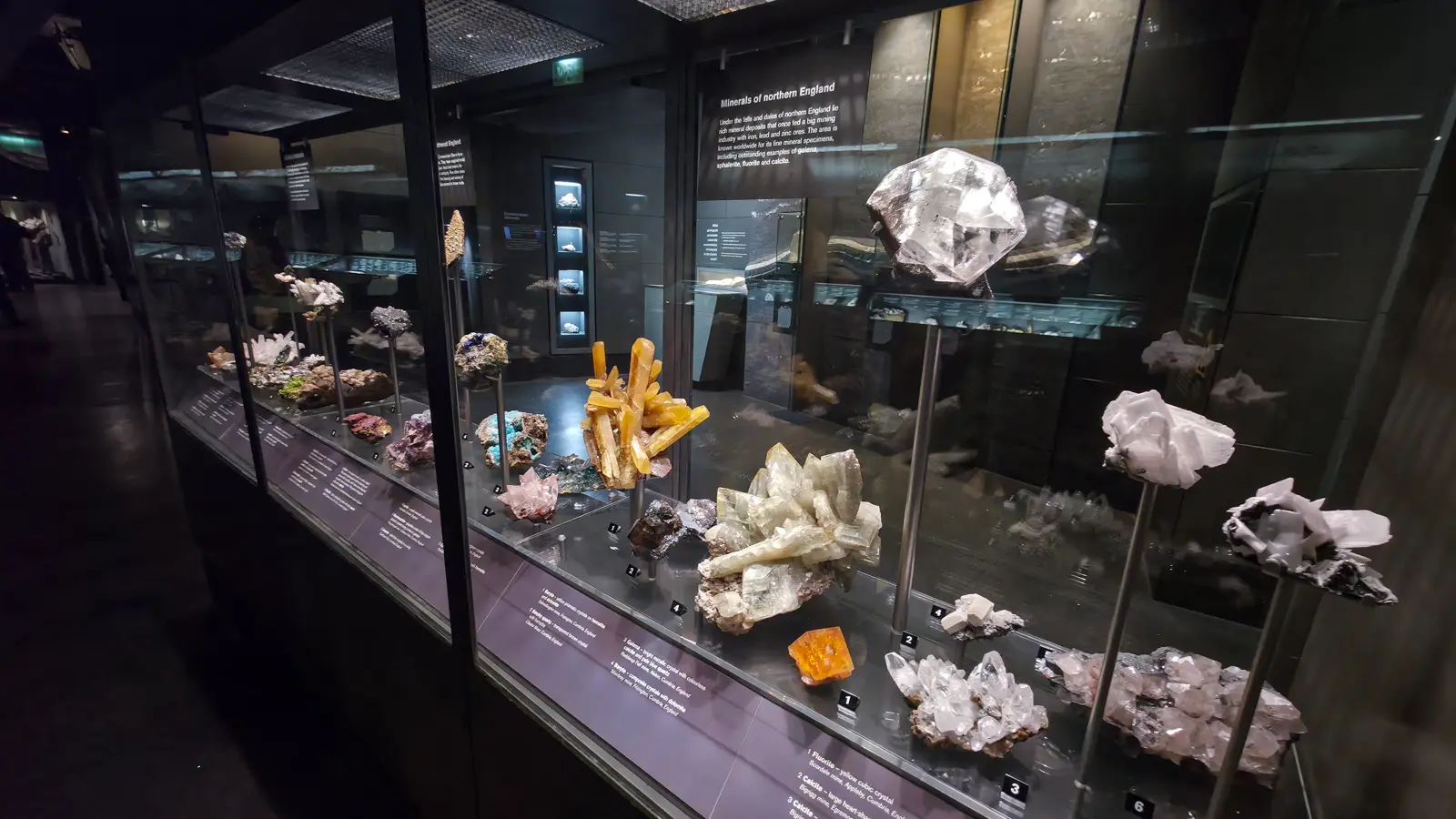 The Minerals Gallery at the Natural History Museum showing the Minerals of Northern England case, with crystal specimens on metal pedestals including a large quartz, calcite clusters, and orange fluorite, against a dark slate background