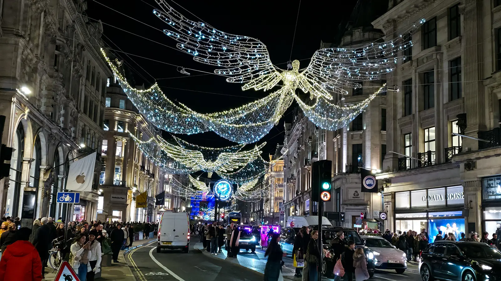 Illuminated white-and-gold flying angel light installations suspended above Regent Street in London during the Christmas season, with crowds on the sidewalks and traffic below