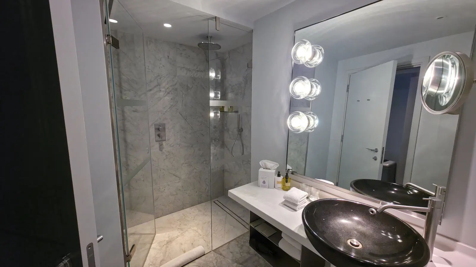 The Standard Room bathroom at Sea Containers London, with white Carrara marble walls and floor, a glass-walled walk-in rainfall shower, a dark stone vessel sink shaped like a polished pebble on a white marble vanity, a large mirror with three round Hollywood-style bulbs running down the left side, and a small porthole-shaped magnifying mirror on the right wall
