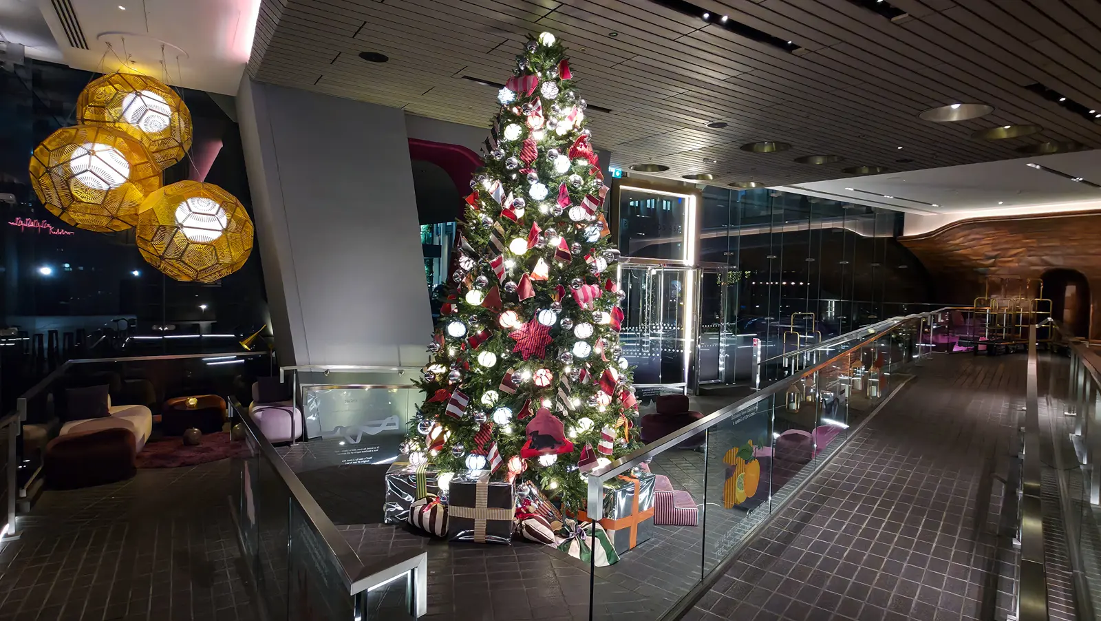 The December Christmas tree in the lobby of Sea Containers London, a tall green tree with warm white lights, silver baubles, and red striped ribbon decorations, with Tom Dixon's patterned gold pendant lanterns hanging on the left side of the upper lobby and the hand-beaten copper hull wall curving behind on the right