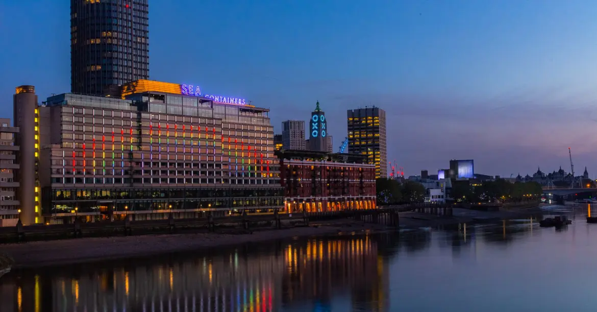 Sea Containers London viewed from across the Thames at twilight, with the multicoloured vertical neon strips lit up across the 16-storey Brutalist concrete façade, the blue Sea Containers rooftop neon signage, the OXO Tower with its iconic blue O-X-O neon to the right, and the river reflections shimmering across the foreground