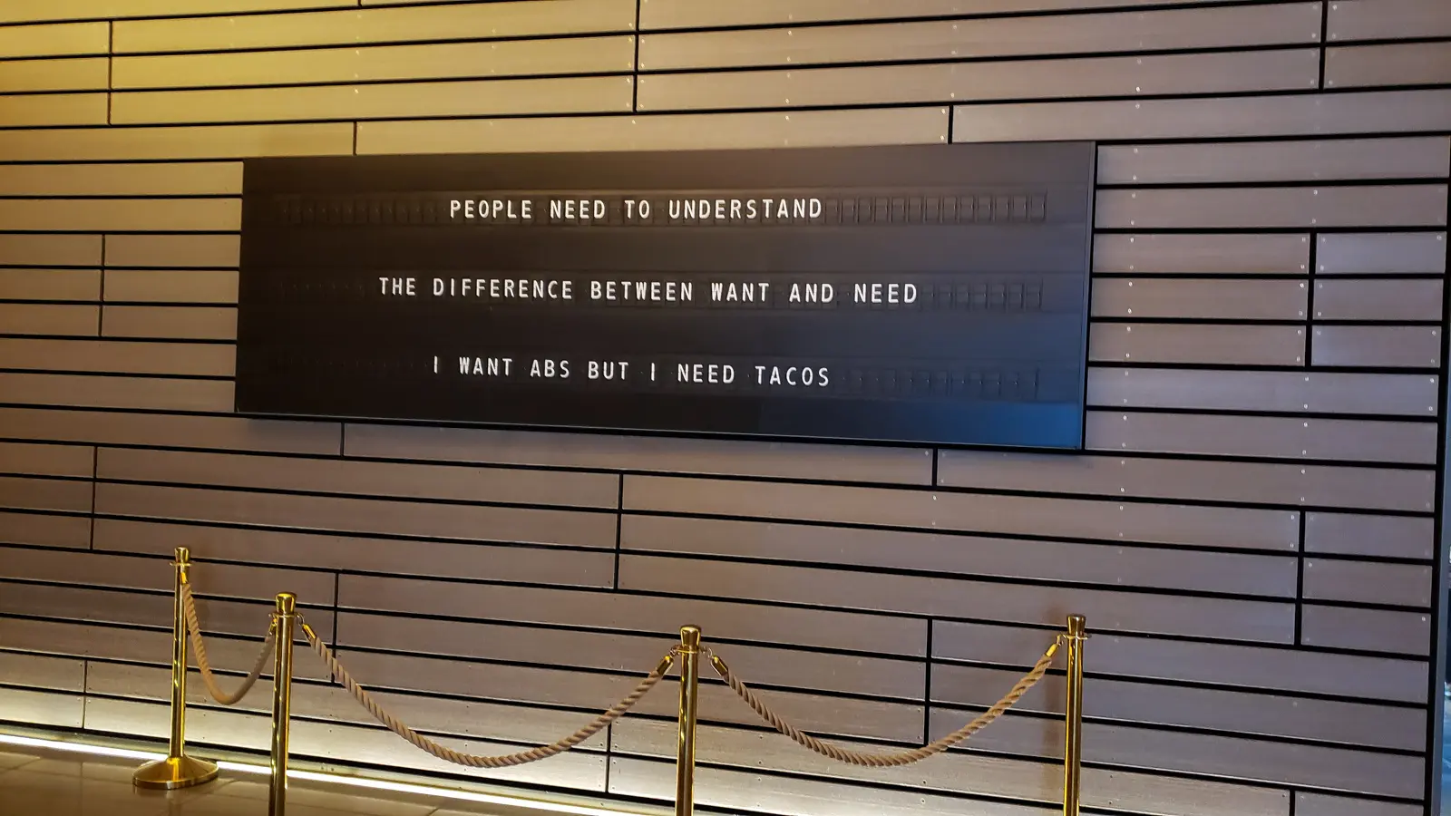 One of Sea Containers London's mechanical split-flap flip-board signs in the lobby, reading PEOPLE NEED TO UNDERSTAND THE DIFFERENCE BETWEEN WANT AND NEED I WANT ABS BUT I NEED TACOS, mounted on a horizontal wood-slatted wall with brass stanchions and a rope barrier in front