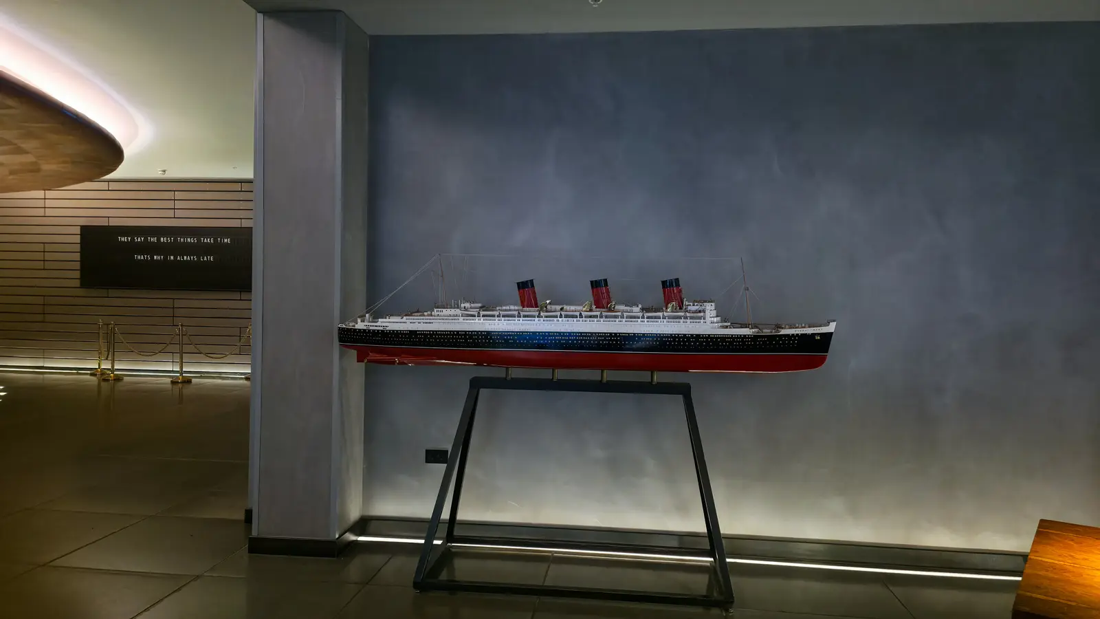 A detailed scale model of the RMS Queen Mary in the lobby of Sea Containers London, in full Cunard livery with a black hull, red waterline, white superstructure, and three iconic red-and-black funnels, mounted on a tall black metal stand against a soft grey painted backdrop wall, with a flip-board sign and brass-stanchion rope barrier visible on the adjacent wood-panelled wall to the left
