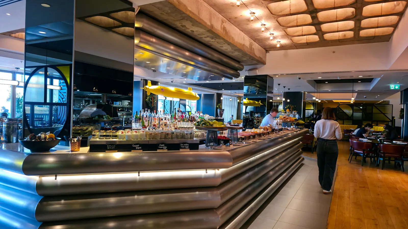 The Sea Containers Restaurant breakfast room with a brushed metal buffet counter in the foreground holding fruit, yogurt, and breakfast items, a yellow submarine model with a small American flag suspended overhead, and the dining area visible in the background with leather chairs on wood plank flooring