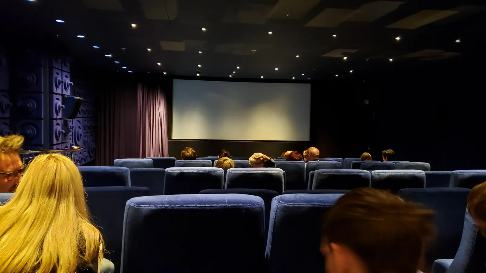 The 61-seat screening room at Sea Containers London, with rows of deep blue velvet seats viewing a white screen, an audience silhouetted in the dim cinema, a brass handrail running along the left aisle, star-like point lights overhead on the dark ceiling, and a repeating concave-dome porthole motif on the left side wall