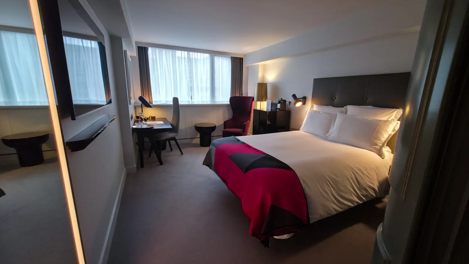 A Standard Room at Sea Containers London showing a queen bed with crisp white linens and a geometric pink-and-black throw at the foot, a dark grey padded headboard, a burgundy velvet wingback armchair in the corner, a polished black writing desk and chair, a tall mirror reflecting the room on the left closet door, floor-to-ceiling windows with sheer curtains, and a light grey carpet