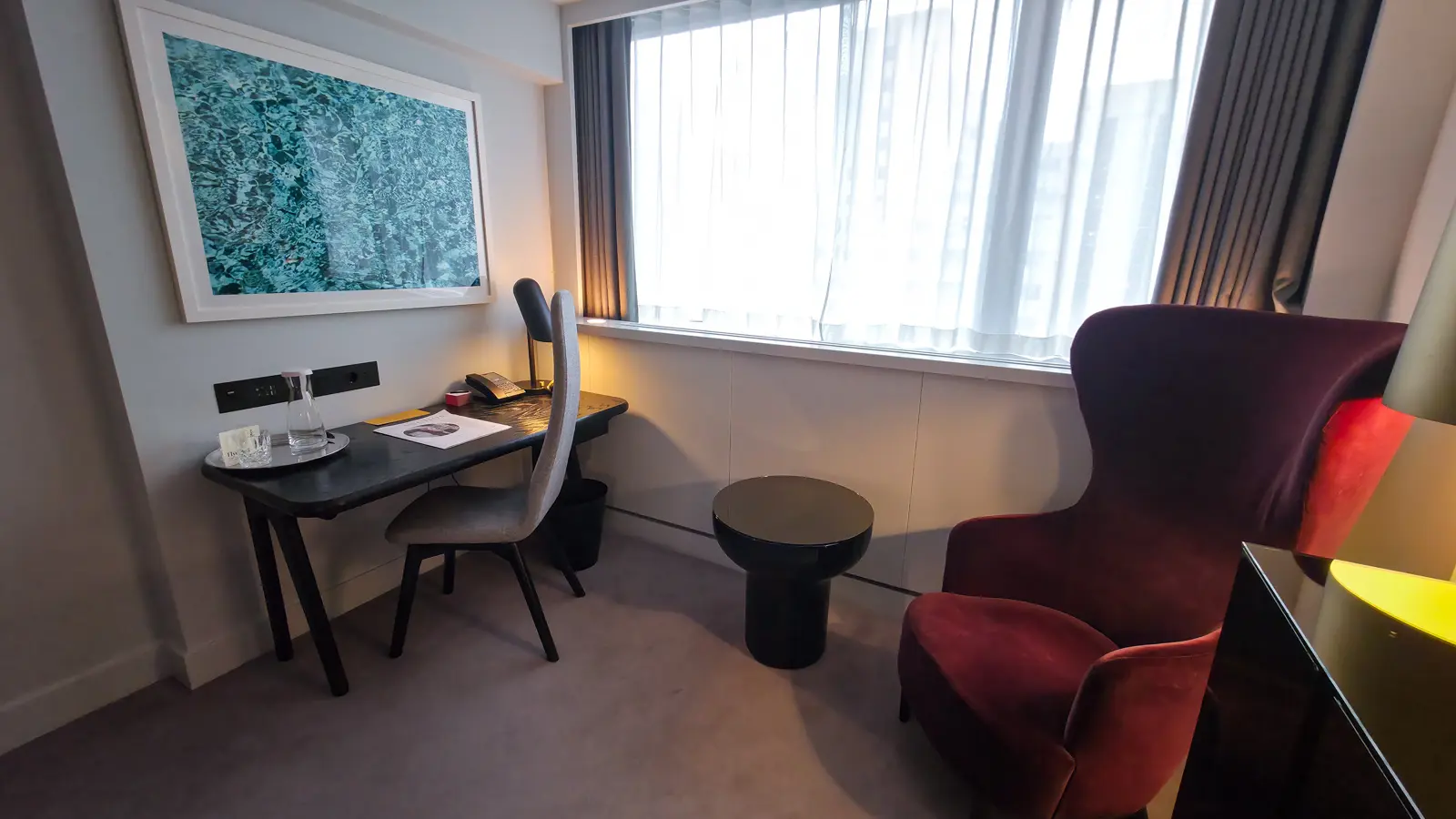 The desk and seating area of a Standard Room at Sea Containers London, with a large framed turquoise water photograph hanging above a polished black writing desk, a modern grey-back desk chair, a glass carafe of water on the desk, a black drum-shaped side table, a burgundy velvet wingback armchair, sheer curtains over a floor-to-ceiling window, and a warm yellow glow from a slim lamp visible at the right edge