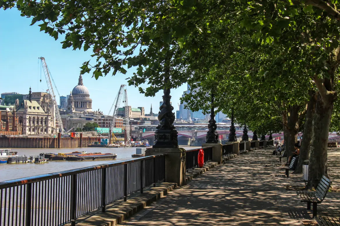 The Queen's Walk promenade along the South Bank in summer, looking northeast toward St Paul's Cathedral and the red iron arches of Blackfriars Railway Bridge. The plane trees and ornate cast-iron lamp posts continue along this stretch of riverside, which runs east from Lambeth Bridge past County Hall, the London Eye, the Southbank Centre, and the National Theatre toward Tower Bridge