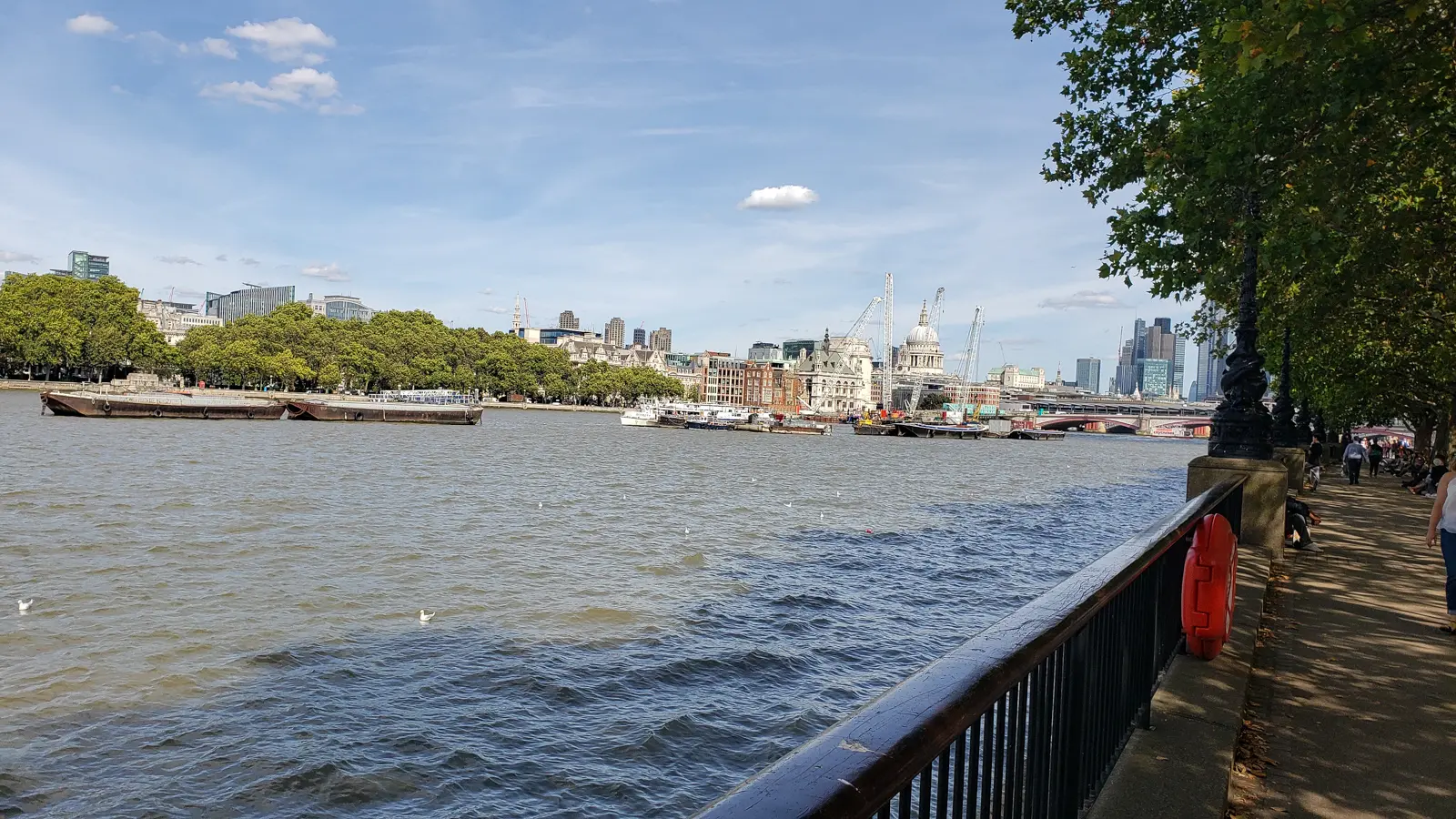 View from the South Bank promenade overlooking the River Thames in London, with boats moving along the river and the dome of St Paul's Cathedral visible in the distance