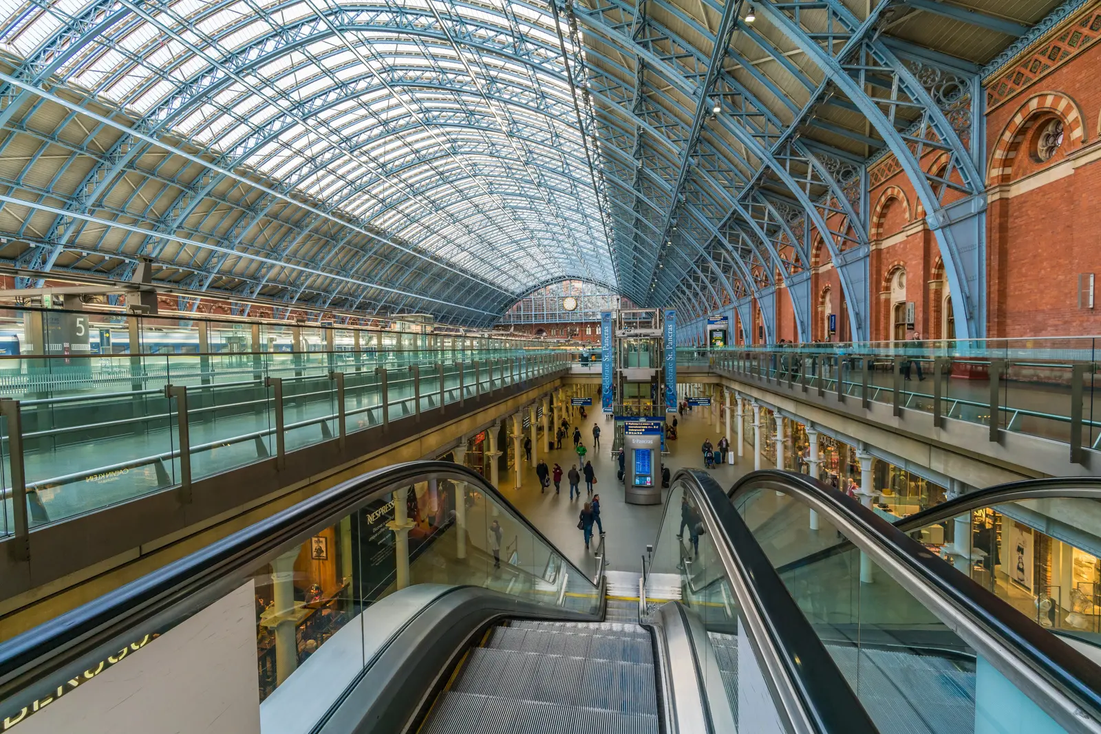 Interior of St Pancras International station in London showing William Henry Barlow's 1868 arched glass-and-iron train shed roof, the red brick walls, escalators to the upper concourse, and shopfronts at platform level