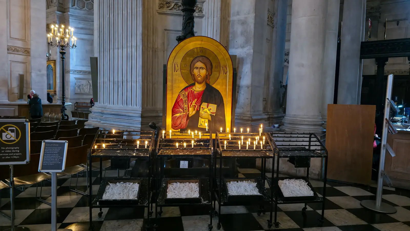 Byzantine-style icon of Christ Pantocrator in the nave of St Paul's Cathedral, holding the Gospel and raising his right hand in blessing, with votive candles burning on tiered black iron stands in front