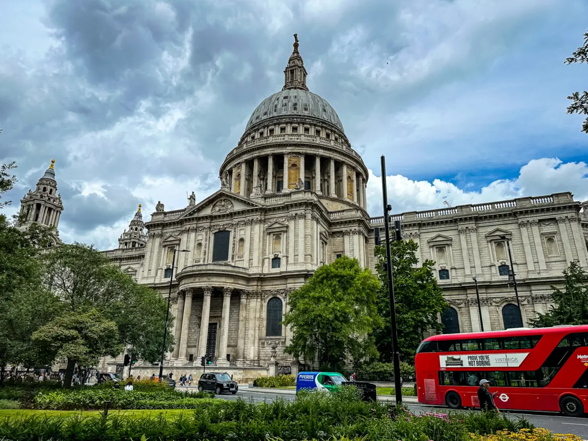 The dome of St Paul's Cathedral viewed from the south side in summer, with a red London double-decker bus passing in the foreground beneath fast-moving cumulus clouds