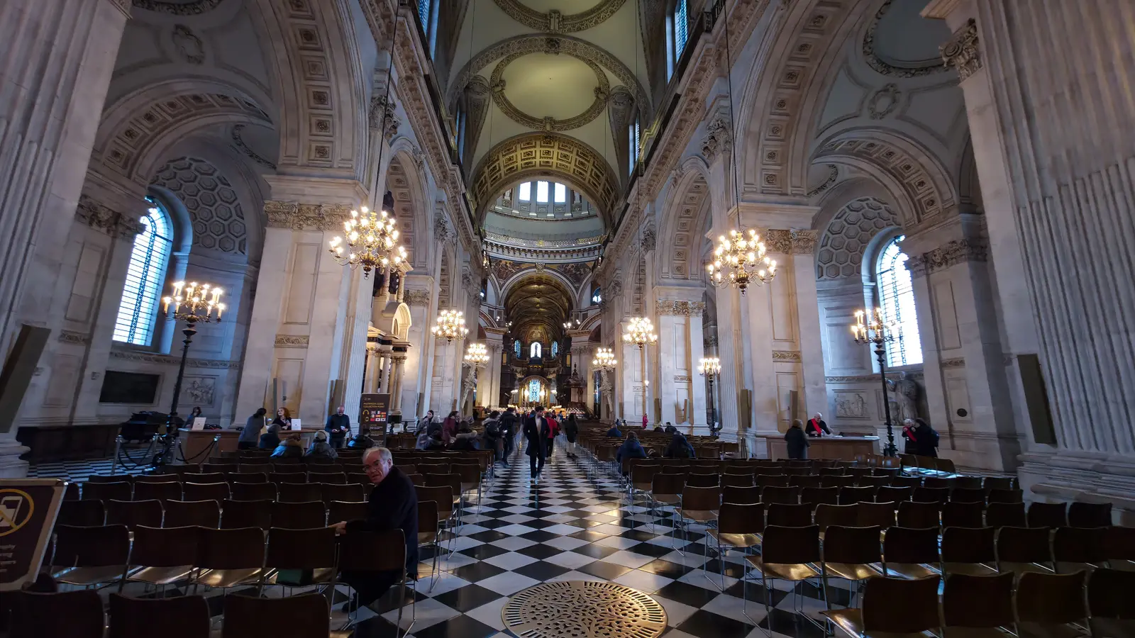 The nave of St Paul's Cathedral looking east toward the choir and dome crossing, with chequered black-and-white marble floor, ornate crystal chandeliers, and rows of wooden chairs