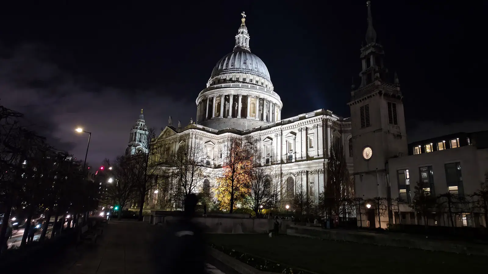 St Paul's Cathedral floodlit at night in December, with the illuminated dome and lantern rising above bare winter trees and the dimly-lit south churchyard