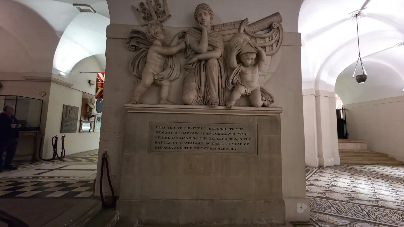 The memorial to Captain John Cooke in the crypt of St Paul's Cathedral, showing a kneeling personification of Britannia flanked by two putti and the stern of a ship, above an inscription marking him as killed commanding HMS Bellerophon at Trafalgar in 1805